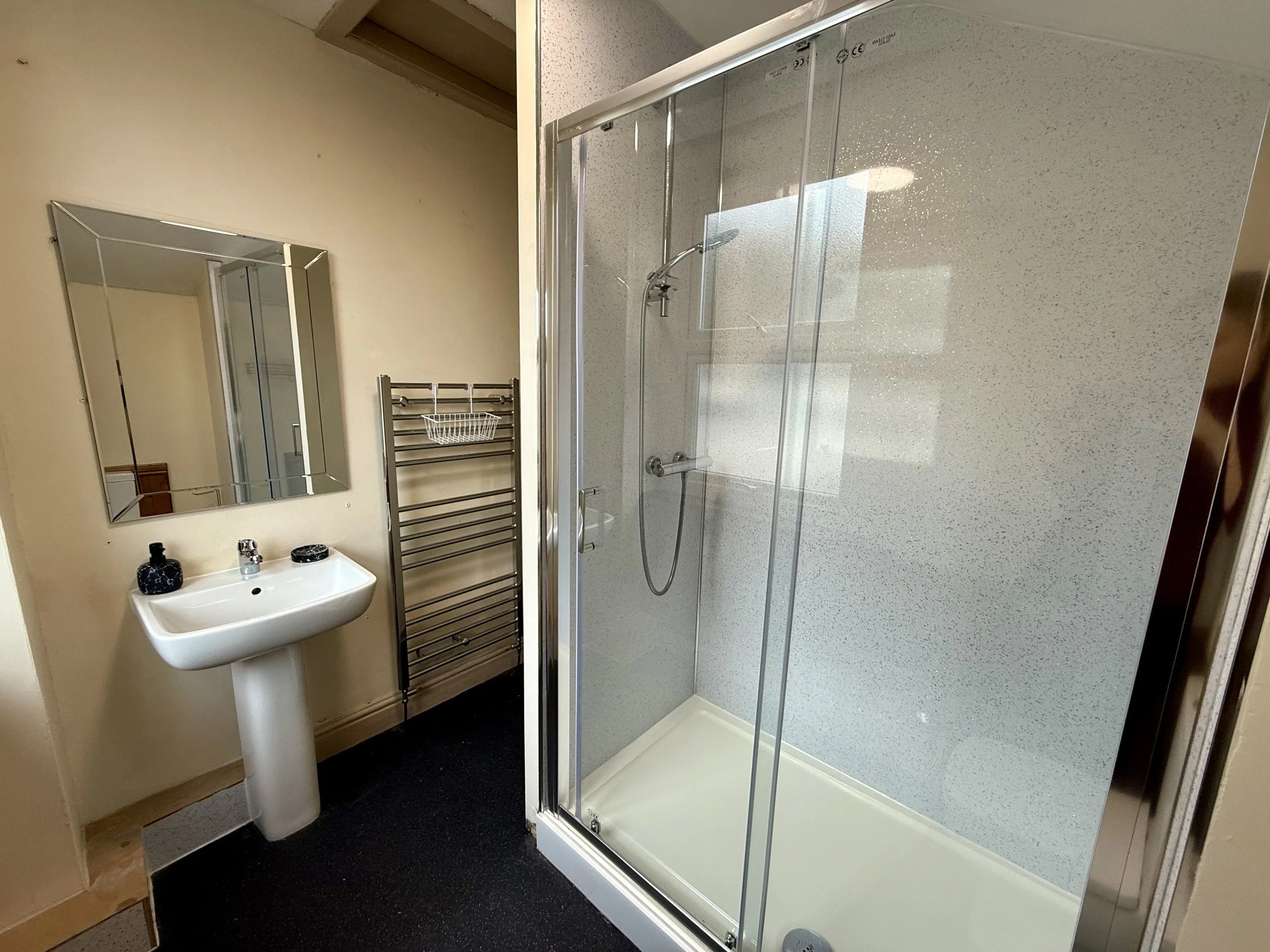 A small bathroom with a white pedestal sink, a wall-mounted mirror, a metal towel rack, and a glass-enclosed shower with a handheld showerhead.