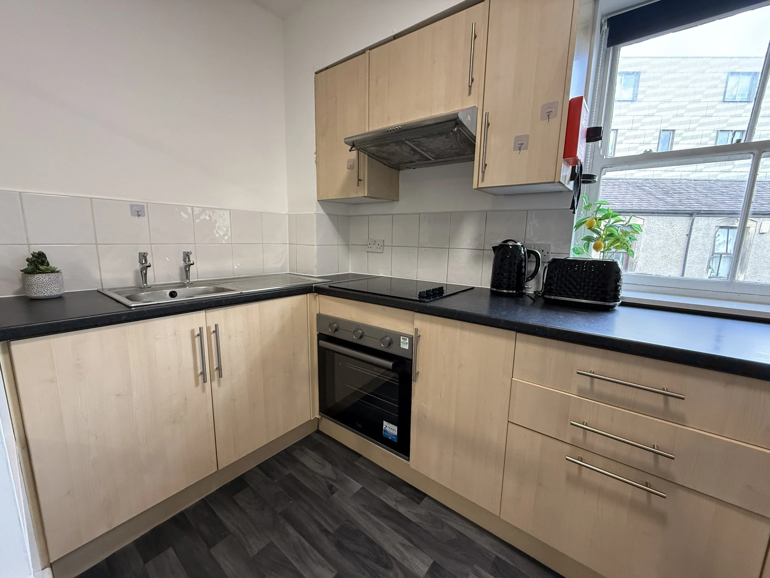 Kitchen with light wood cabinets, black countertops, a window with a view of neighboring buildings, and small appliances including a kettle and toaster on the counter.