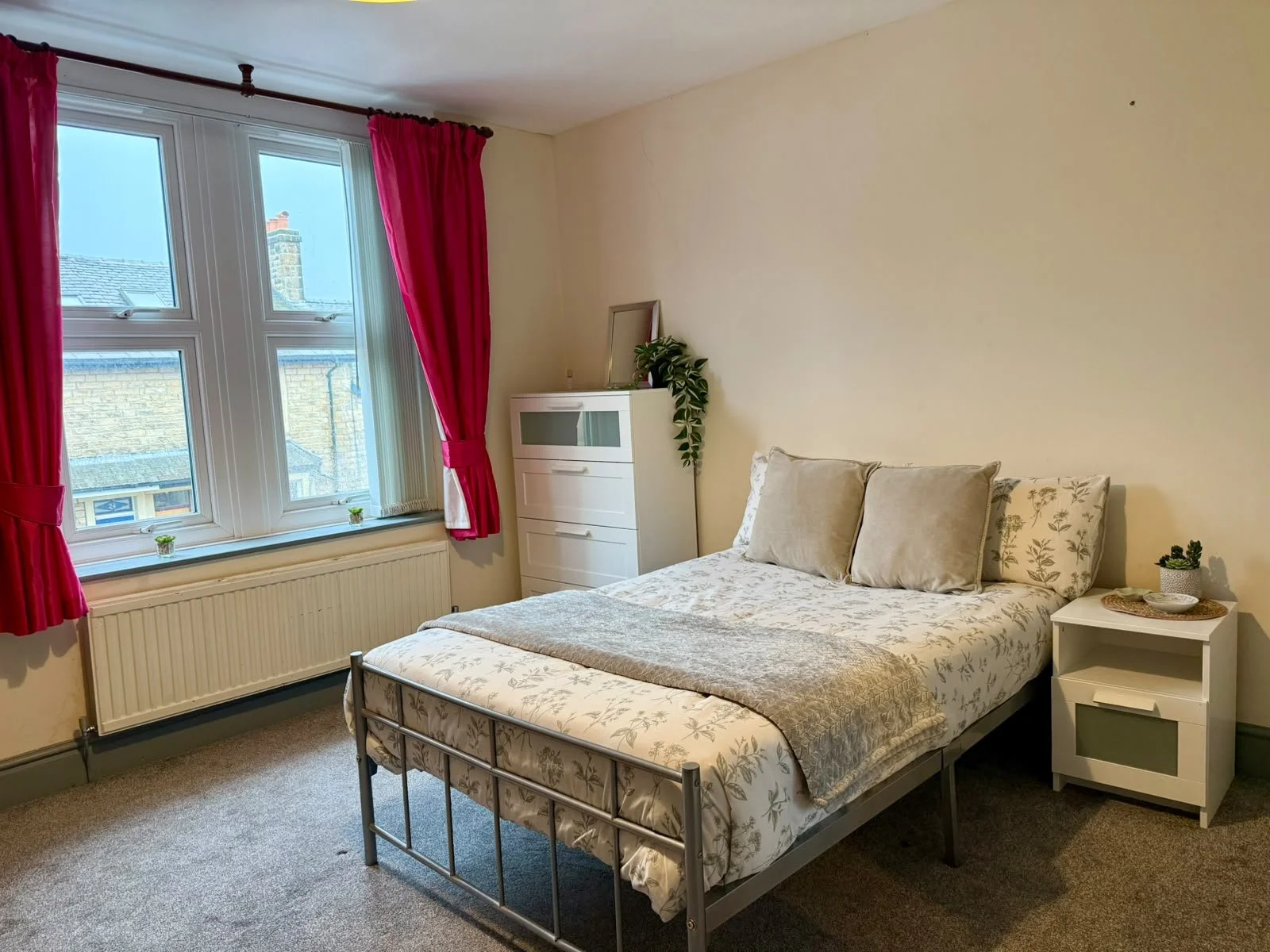 A bedroom with a bed, white floral bedding, beige pillows, a bedside table with a small plant, and a window with red curtains showing rooftops outside.