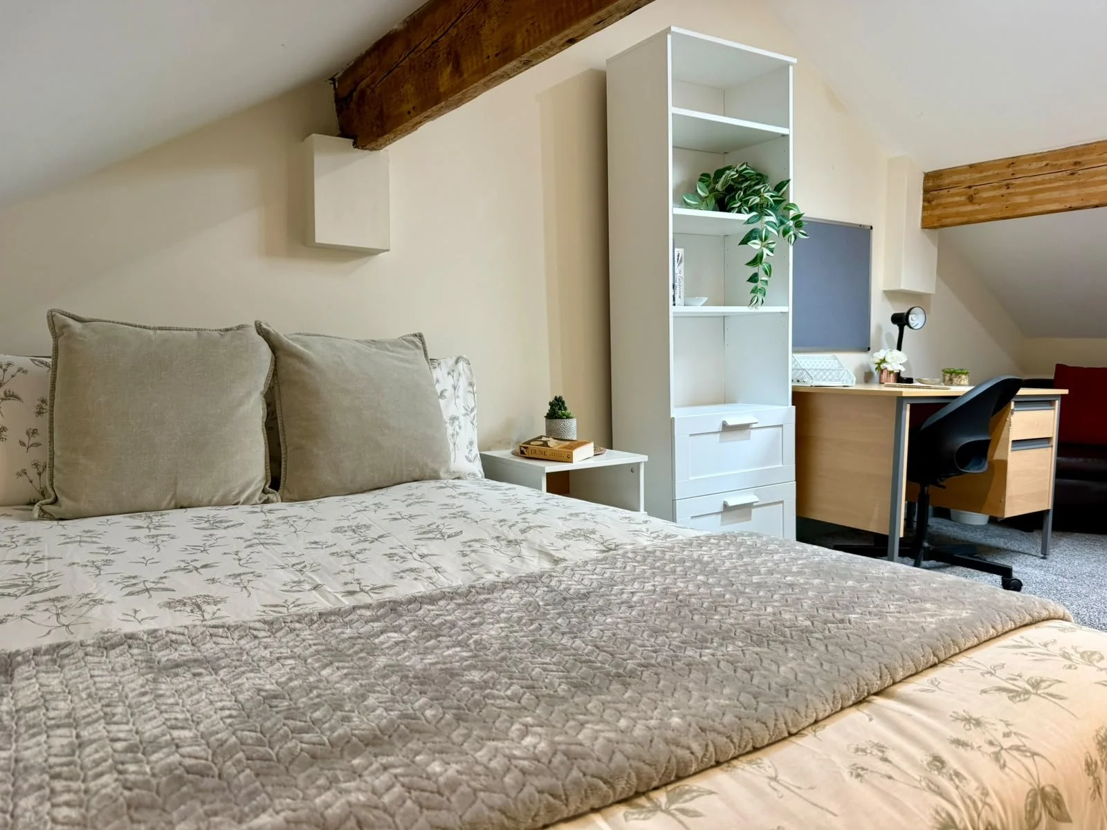 A cozy bedroom with a bed featuring beige pillows and a patterned bedspread, a small side table with a plant and books, a white bookshelf, a desk with a black chair, and wooden ceiling beams.