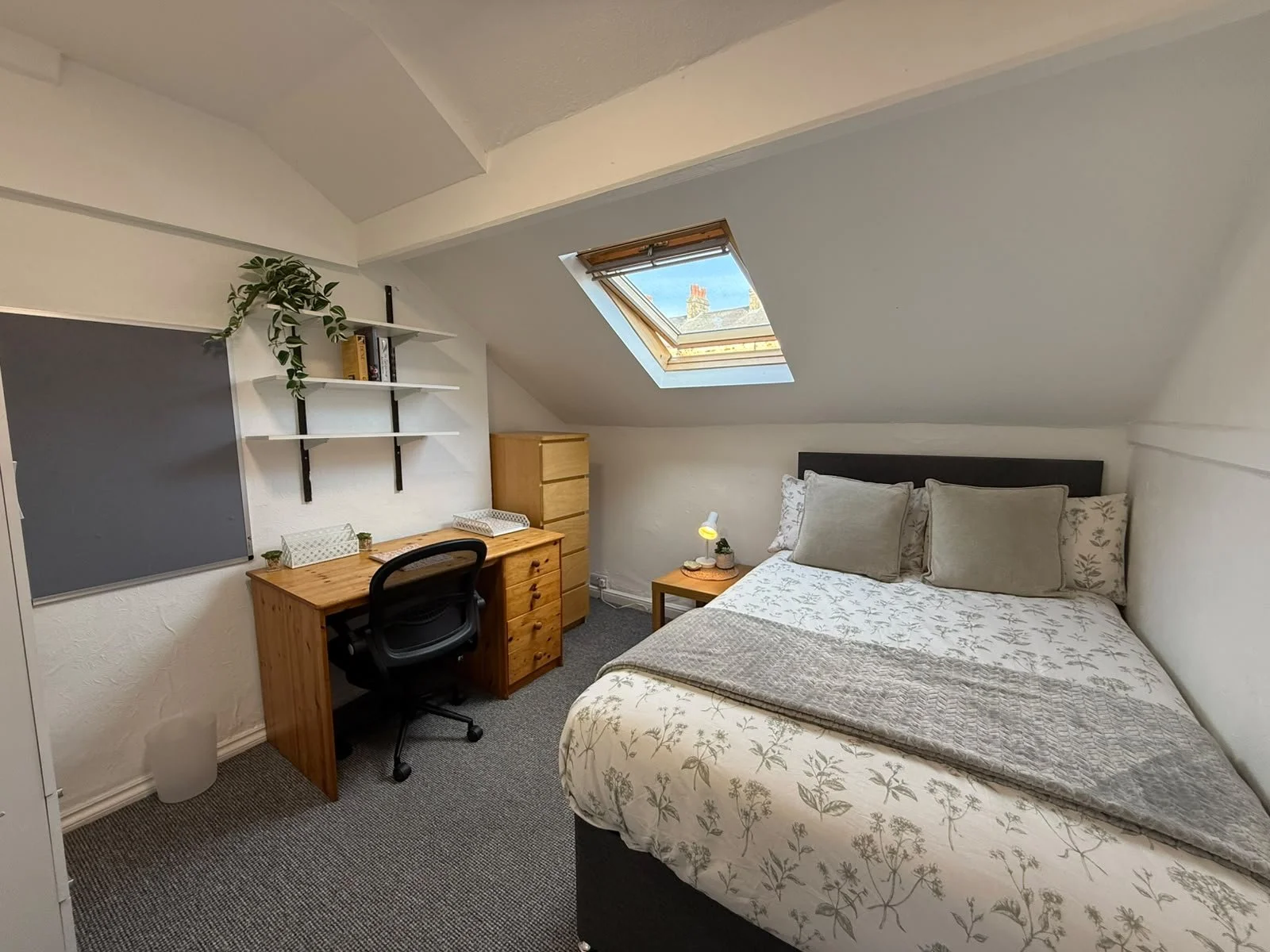 A cozy bedroom with a sloped ceiling and a skylight window. There's a bed with light-colored floral bedding, a small wooden nightstand with a lamp and decorative items, a wooden desk with a chair, a small wooden dresser, and wall-mounted shelves with