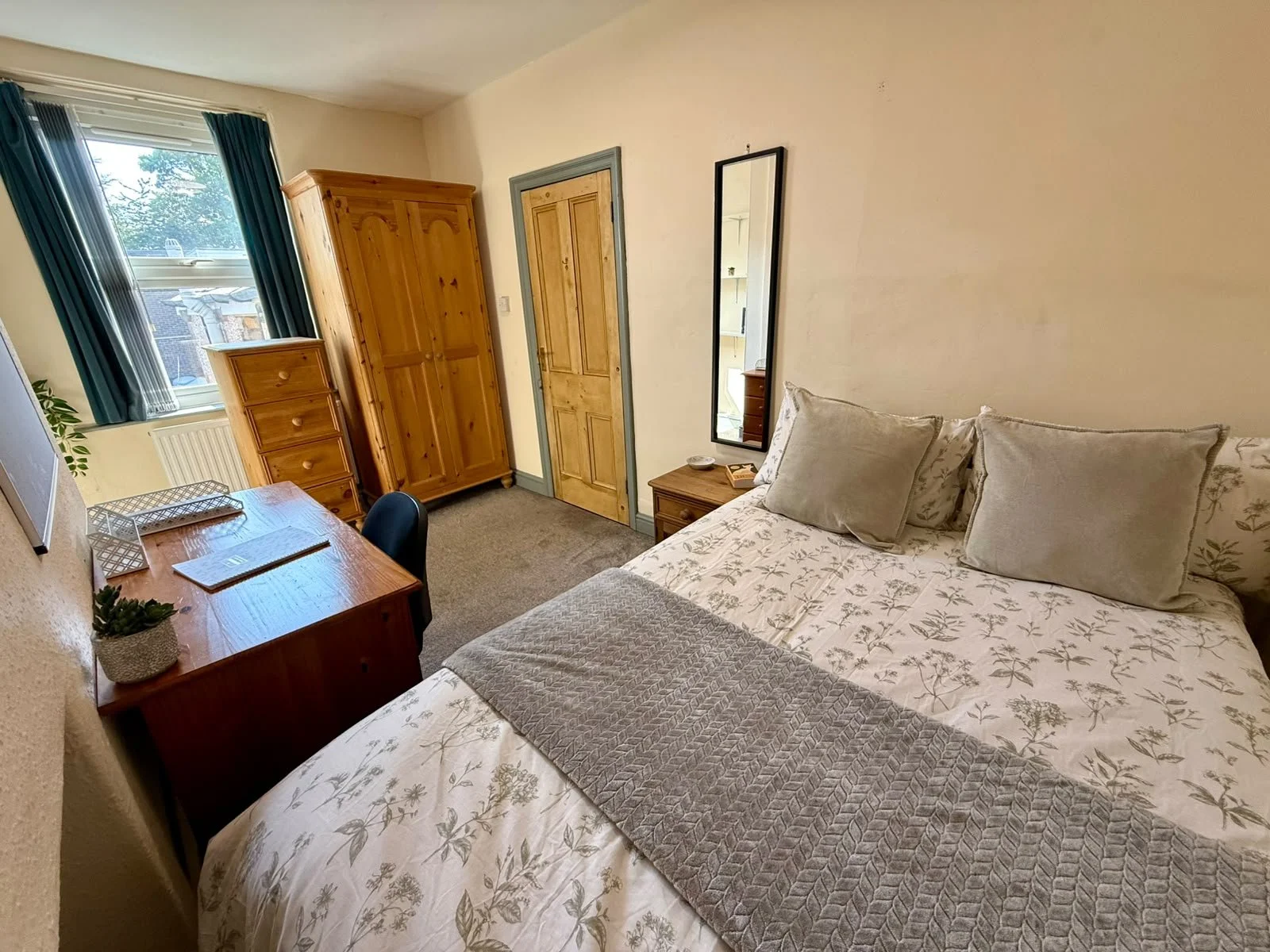 A cozy bedroom with a large bed covered in floral bedding and pillows. A small wooden nightstand with a dish and book, a tall mirror, and a wooden wardrobe are visible. A window with blue curtains lets in natural light, and there is a desk with a cha