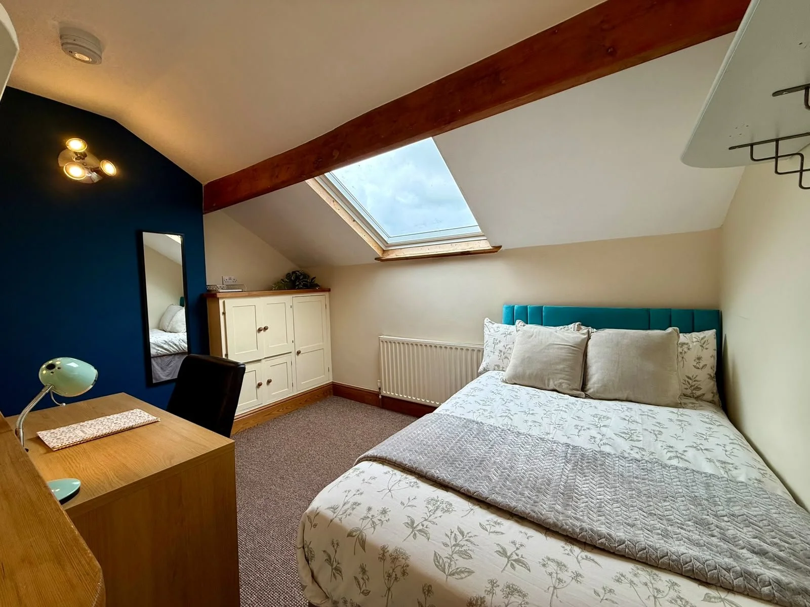 A cozy bedroom with a sloped ceiling and a skylight window showing a cloudy sky. The room features a bed with a light-colored floral duvet and multiple pillows, a small wooden desk with a vintage green desk lamp, a black chair, a white cabinet, and a