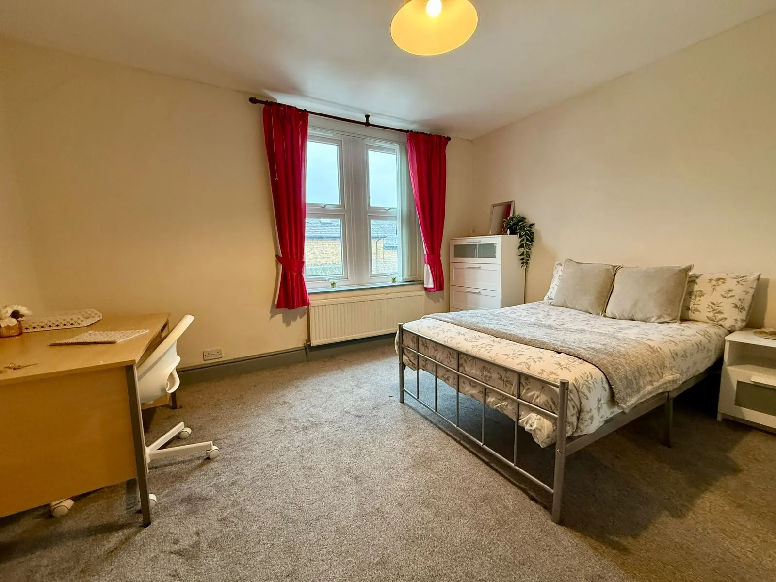 A bedroom with a window, beige walls, a bed with pillows and a floral bedspread, a white dresser, a small white nightstand, a wooden desk with a white chair, red curtains, and a ceiling light.