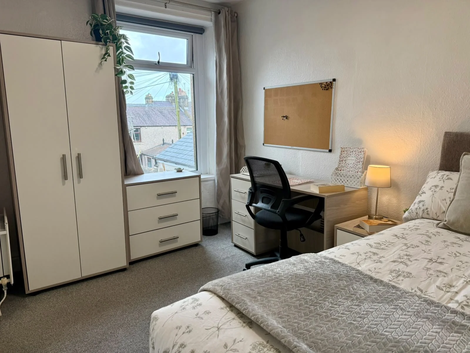 A cozy dorm room with a bed, a desk, a black office chair, a white nightstand with a lamp, a white wardrobe, and a window with beige curtains. There is a bulletin board on the wall above the desk and a cardboard box on the nightstand. The window show