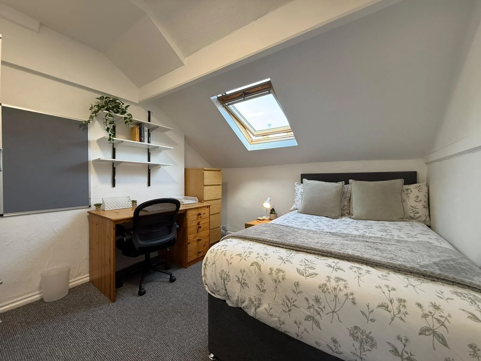 A cozy attic bedroom with a bed, desk, and ceiling skylight, decorated with neutral tones and houseplants.