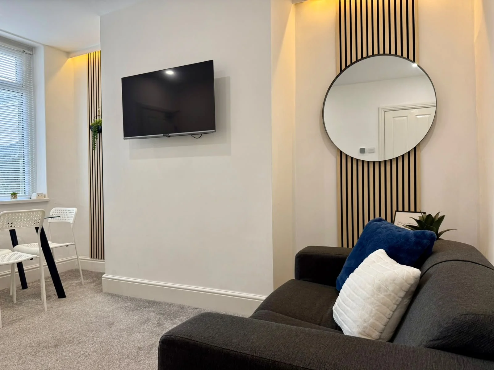 Living room area with a gray sofa with white and blue cushions, a wall-mounted flat-screen TV, a round mirror, white chairs, and a window with blinds, decorated with small potted plants.
