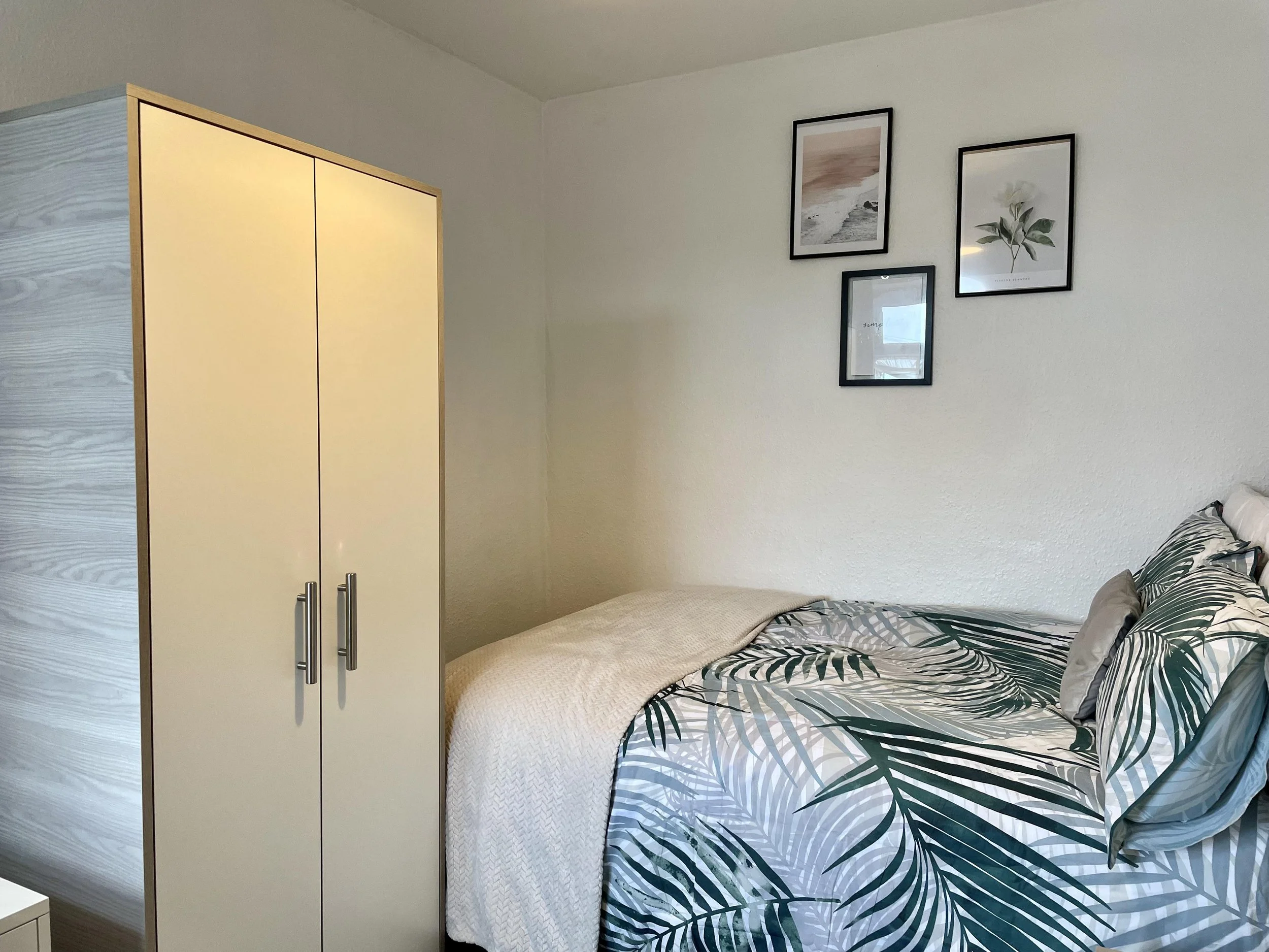 A neatly made bed with tropical leaf-patterned bedding, a cream blanket, and multiple pillows, next to a white wardrobe, with three framed pictures on a white wall.