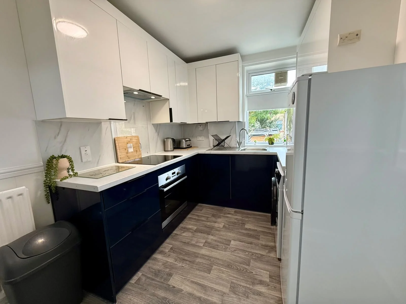 Modern kitchen with white upper cabinets, navy blue lower cabinets, marble backsplash, and a window above the sink looking outside. Appliances include a built-in oven, refrigerator, microwave, and small appliances like a kettle and toaster on counter