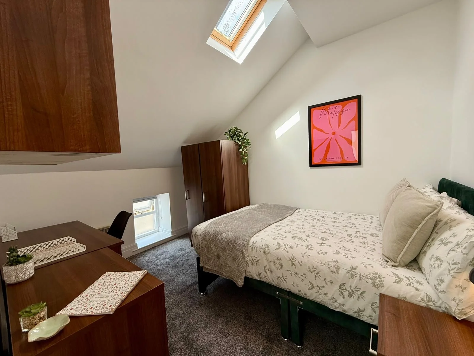 A bedroom with a sloped ceiling, skylight, and small window. Contains a bed with floral patterned bedding, a wooden wardrobe, a framed pink and orange art print, a small desk with decorative items, and a potted plant.