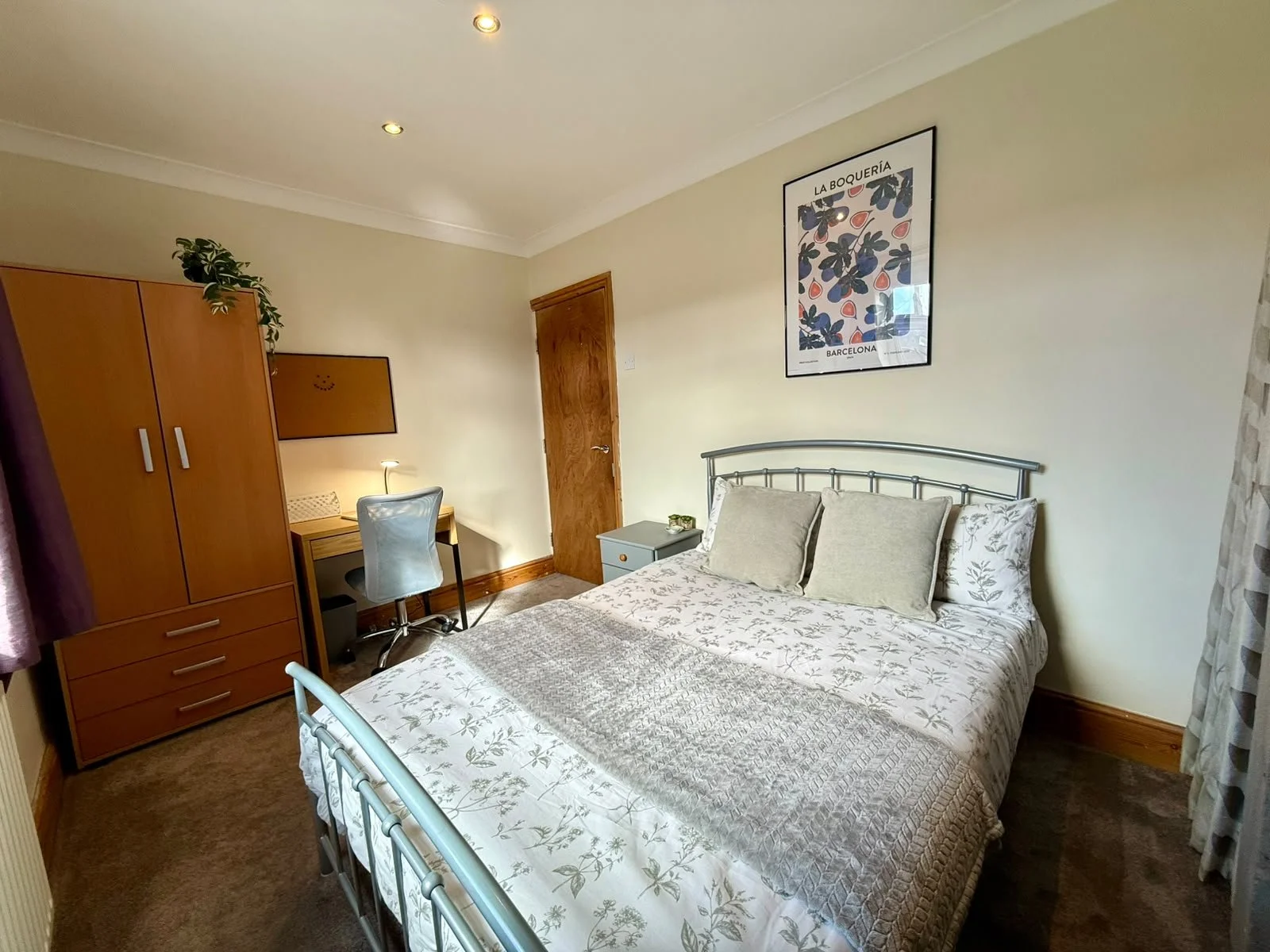 A cozy bedroom with a bed, pillows, a nightstand, a wardrobe, a small desk with a chair and lamp, and framed artwork on the wall.