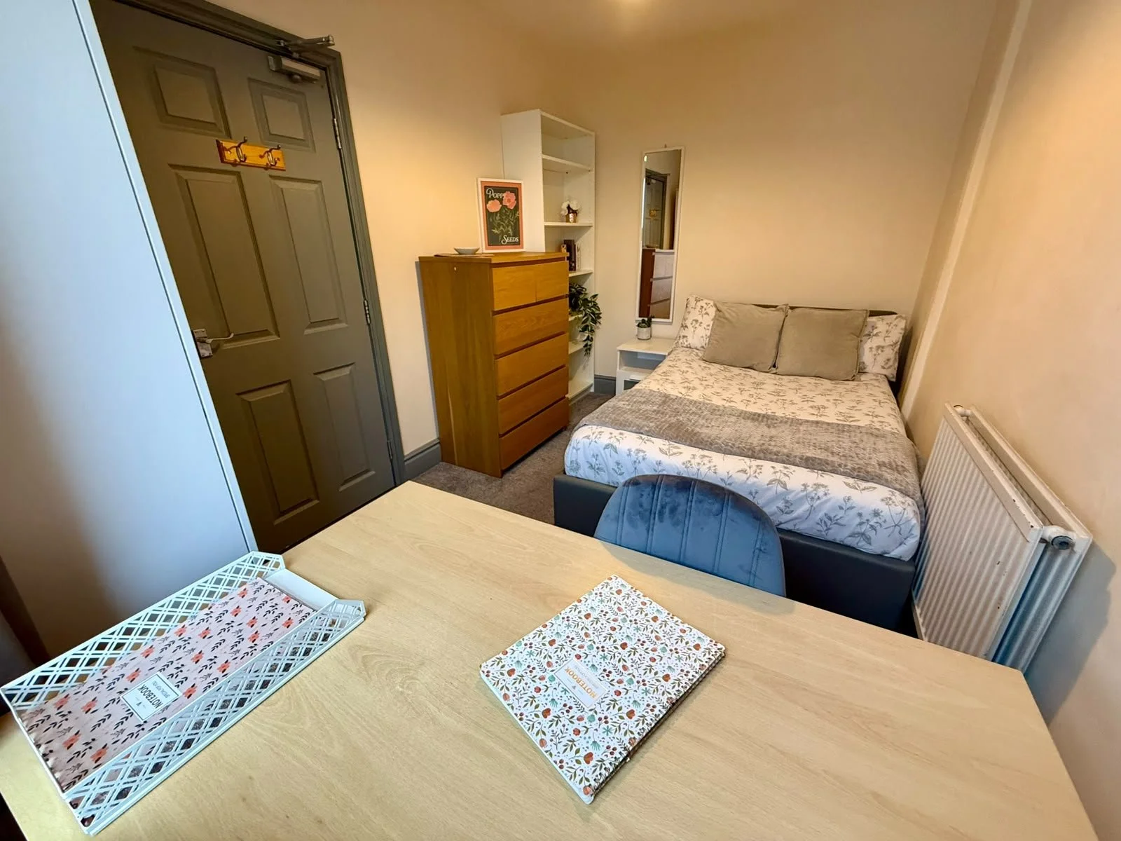 A cozy bedroom with a bed, a white nightstand, and a tall wooden dresser. There is a small mirror and some decorative items on the shelves above the nightstand. The room has a door, a white radiator, and a desk with a chair. The desk has a tray, a fo