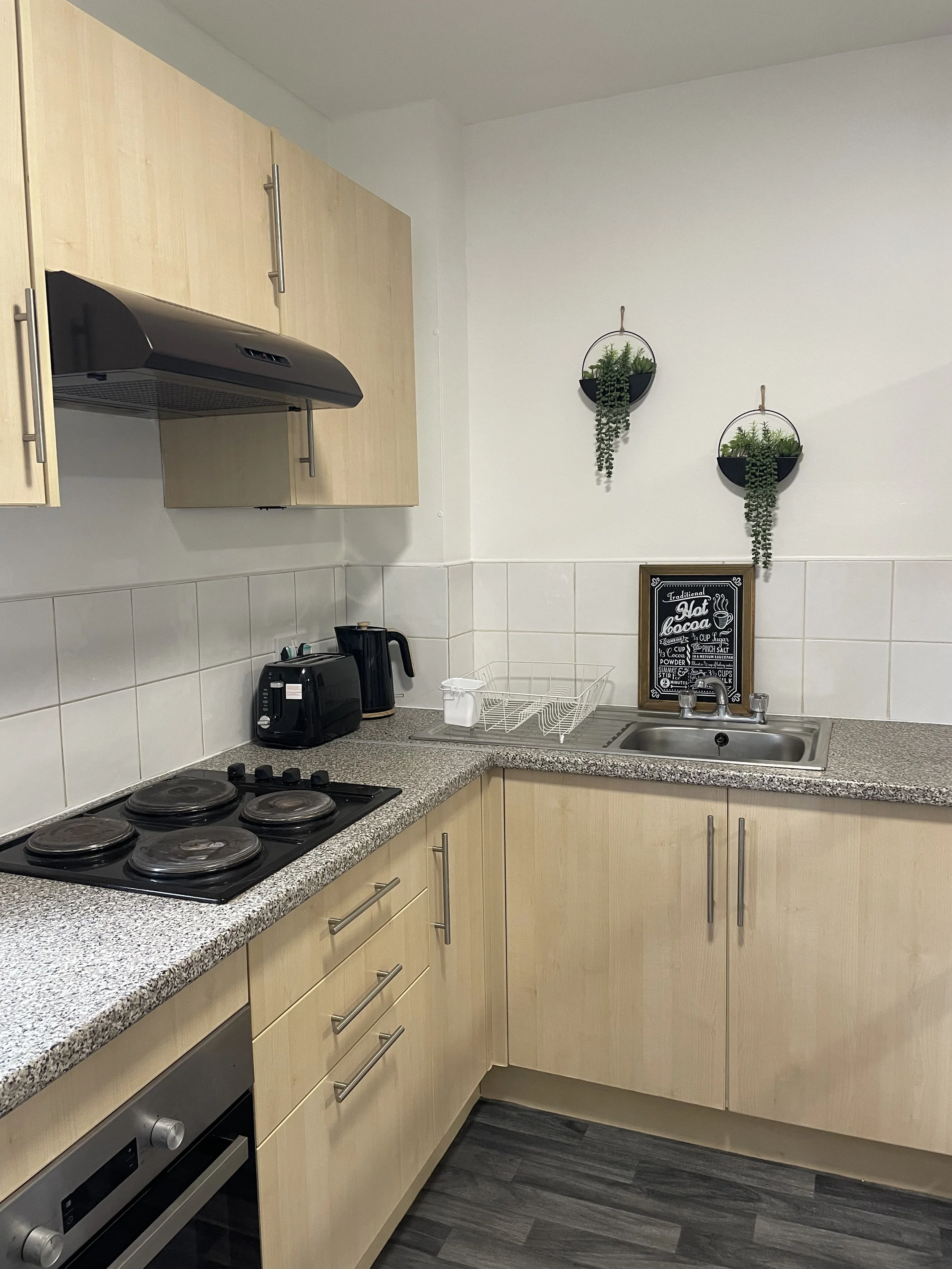 Kitchen with wooden cabinets, a black stove with four burners, a countertop with a toaster and electric kettle, a dish drying rack, a small framed chalkboard sign, and two hanging planters with greenery on the white wall.