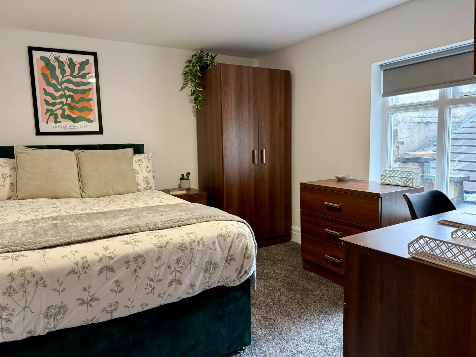 A cozy bedroom with a bed featuring green velvet headboard, floral bedding, and multiple pillows, a framed abstract wall art above, a wooden wardrobe with plant on top, a wooden dresser, and a window with closed blinds overlooking outdoor stairs.