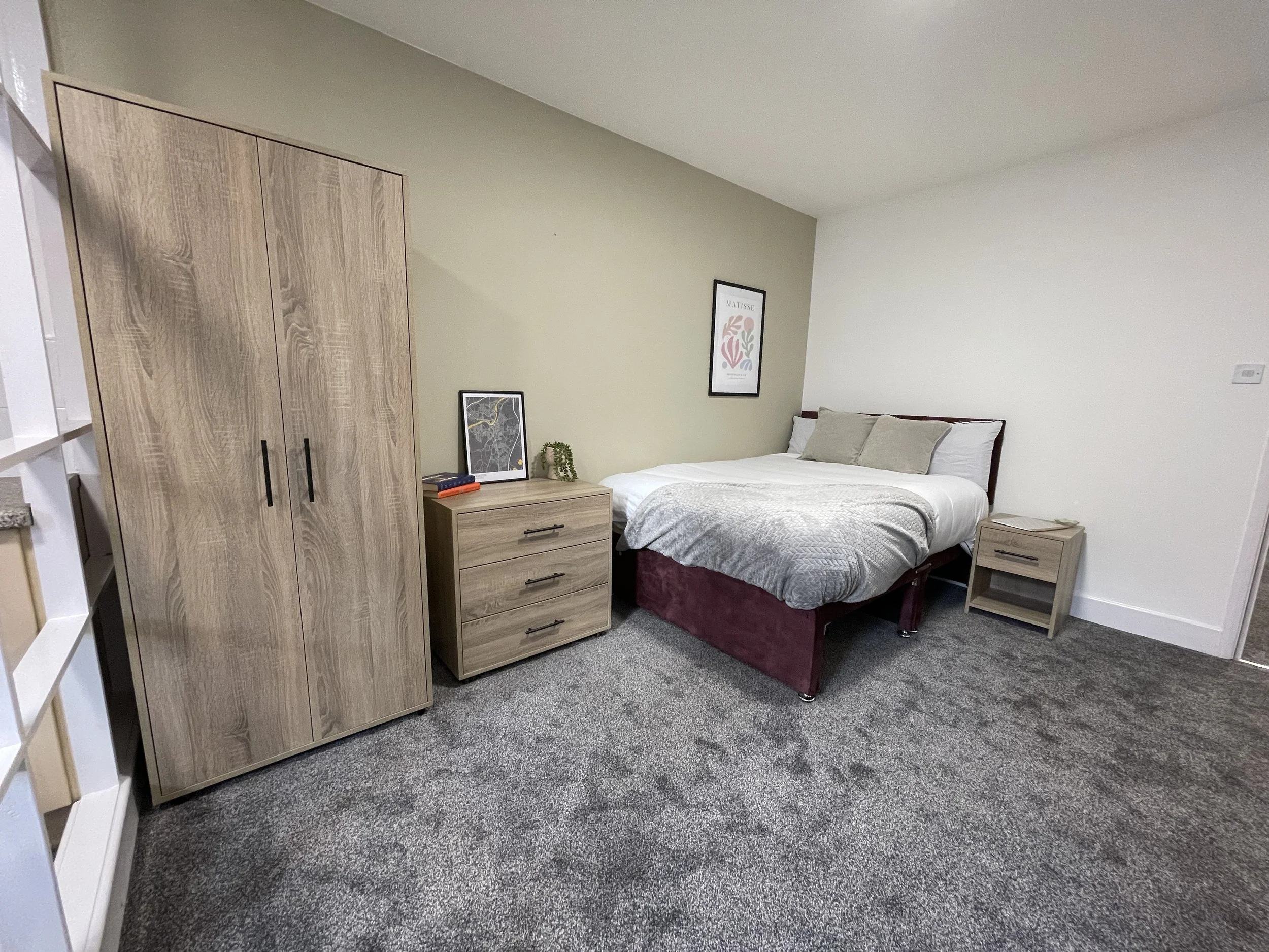 A bedroom with a bed and nightstand, a wardrobe, a dresser with a picture frame and decorative item, and framed art on the wall. The room has neutral colors and carpet flooring.