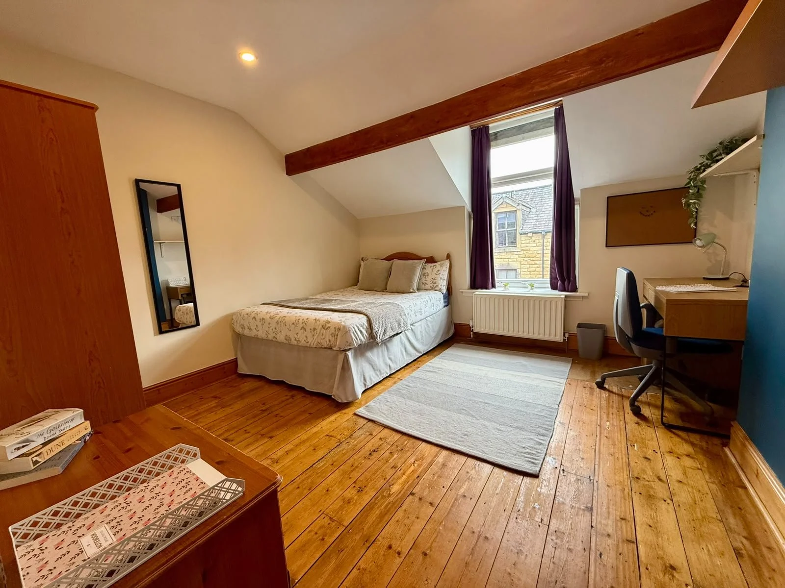 A cozy bedroom with wooden flooring, a bed with floral bedding near a window with purple curtains, a small desk with a chair and lamp, a mirror on the wall, and a bookshelf with books.