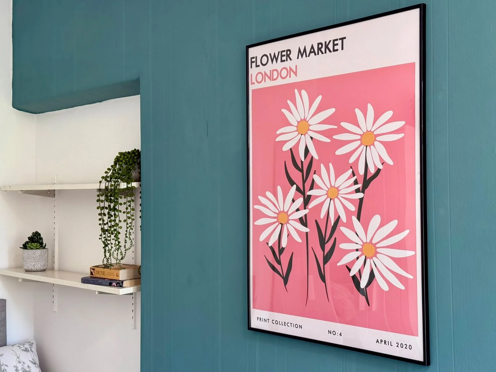 A poster on a teal wall advertising a flower market in London with white daisies on a pink background, titled 'Flower Market London'.