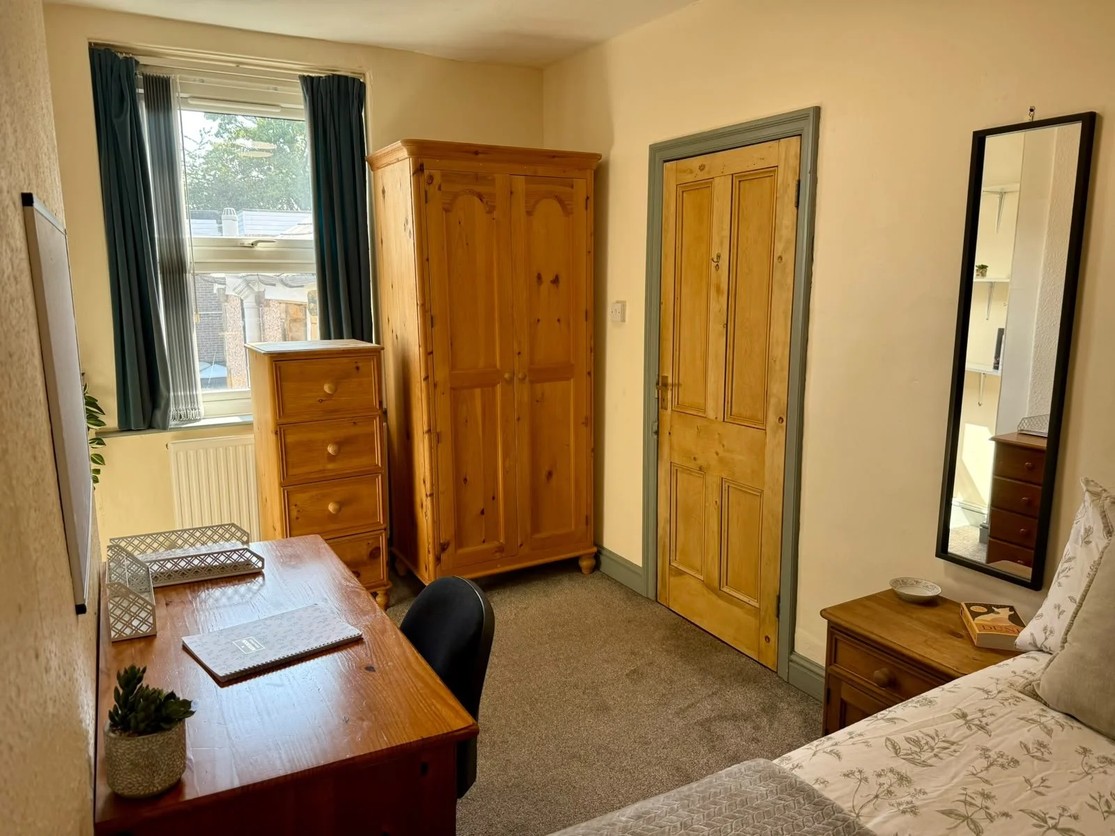 A small bedroom with a wooden desk, a chair, a tall wooden wardrobe, a small wooden chest of drawers, a full-length mirror, a window with curtains, and a bed with patterned bedding.