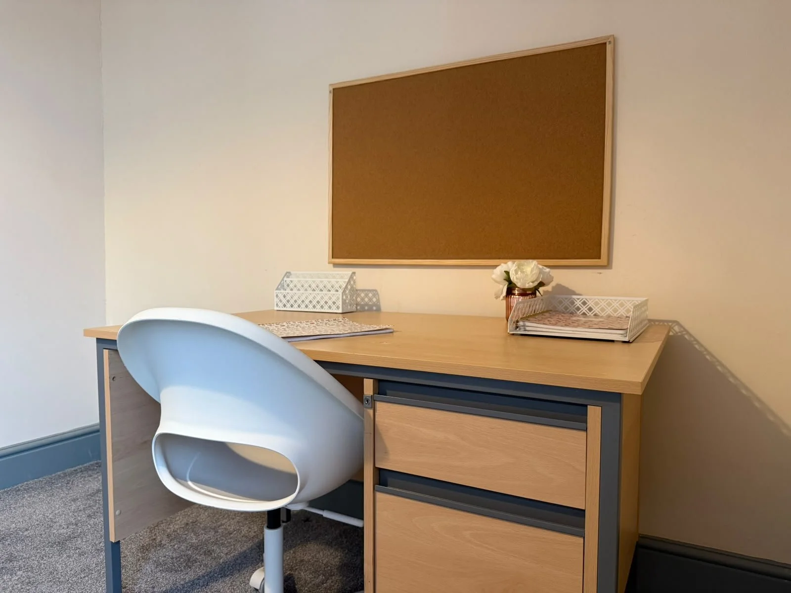 A small office or study room with a wooden desk, a white modern chair, a corkboard on the wall, and a vase with white flowers on the desk. There are also two wire baskets and a file organizer on the desk.