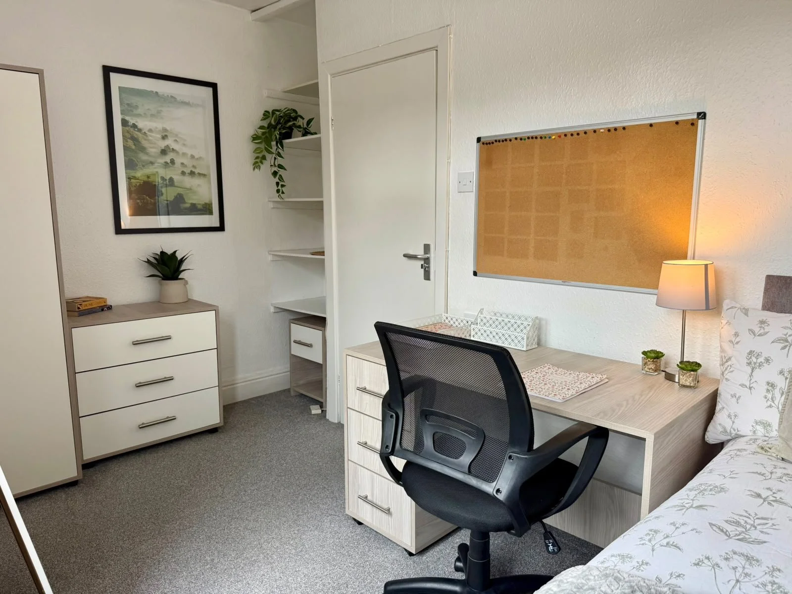 A cozy bedroom with a desk, black ergonomic chair, white drawers, potted plants, wall art, bulletin board, and a bed with floral bedding, warm lighting from a table lamp.