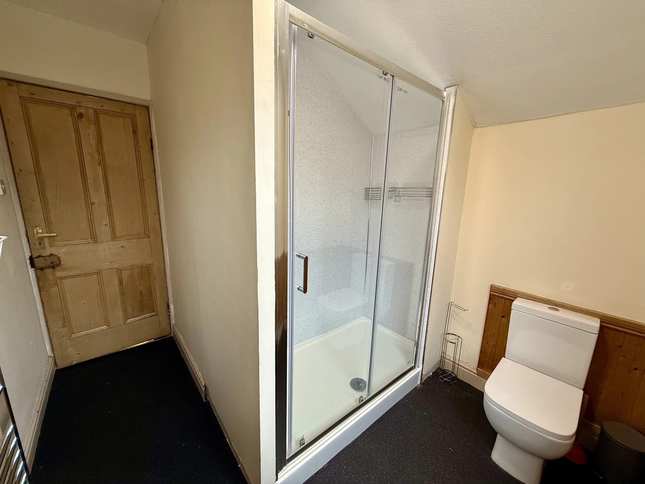Bathroom with wooden door, shower with glass door, toilet, and wood paneling on the wall behind the toilet.
