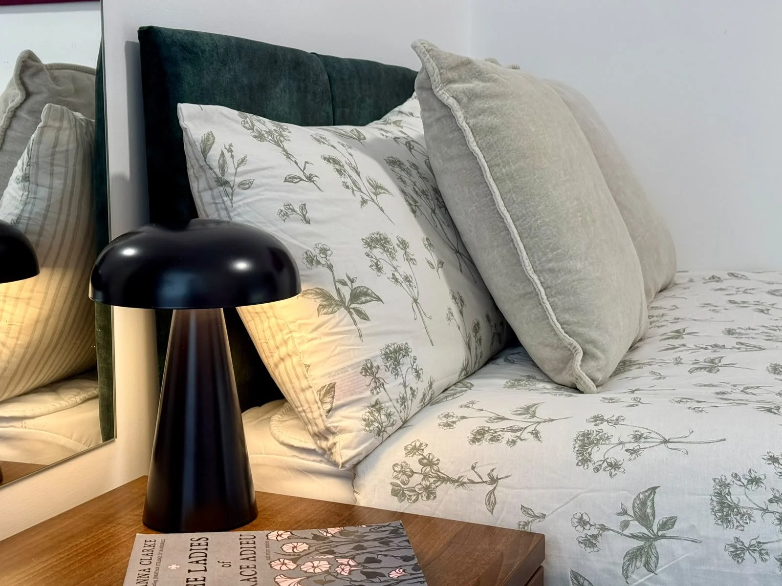 A bed with floral-patterned white and gray bedding, gray pillows, and a dark green headboard, with a black bedside lamp and a mirror on the wall.