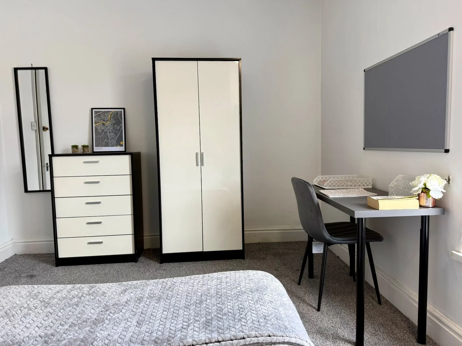 Amidst a tidy, modern bedroom, furniture includes a white dresser with black border, a matching wardrobe, a tall mirror, and a small desk with a chair. The desk has a flower vase, books, and notebooks, and a gray bulletin board hangs above it.