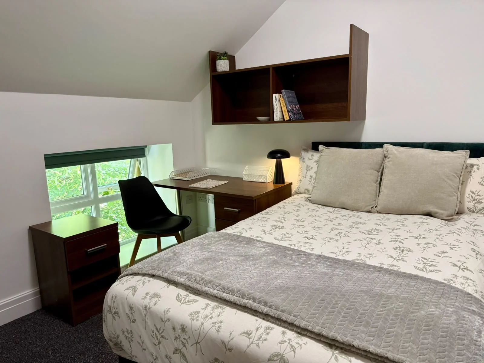 A bedroom with a bed, pillows, window, desk, chair, wall shelf, and a small bedside table with a lamp.
