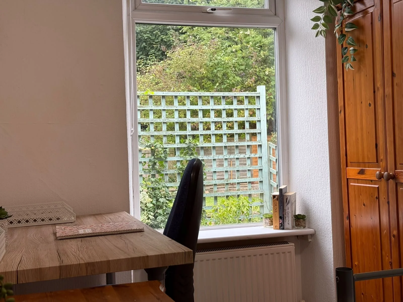 Interior view of a room with a large window overlooking a garden with greenery and a wooden fence. Inside, there is a wooden table with a notebook, a basket, and a small potted plant. To the right, there are wooden cabinets, and on the windowsill, th