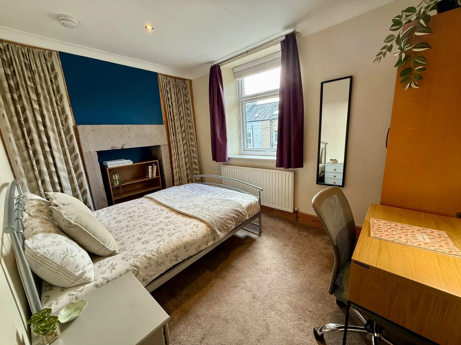 A cozy bedroom with a single bed, window with purple curtains, a wooden desk with a chair, mirror, and a small bookshelf, carpeted floor, and a fireplace mantle on the wall.