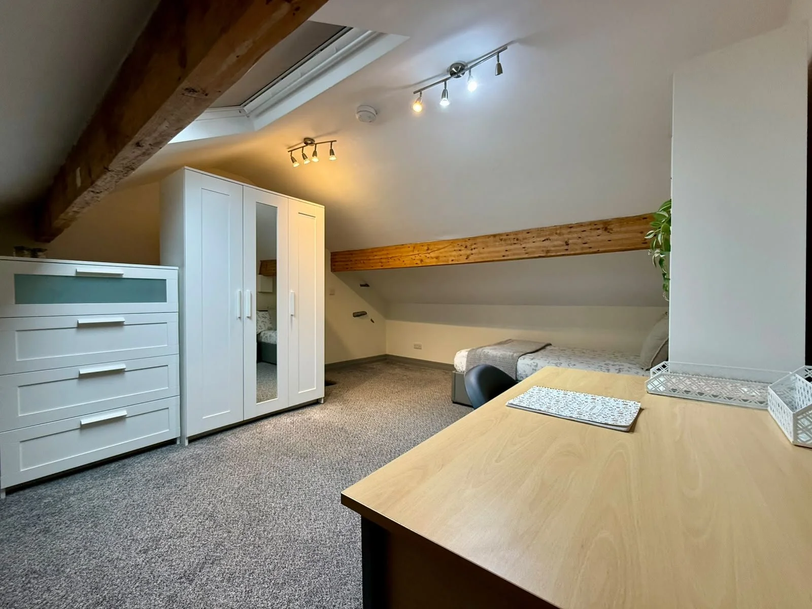 A cozy attic room with sloped ceilings and exposed wooden beams, featuring a white wardrobe, a chest of drawers, a bed, a small desk, and a skylight window with overhead lighting.