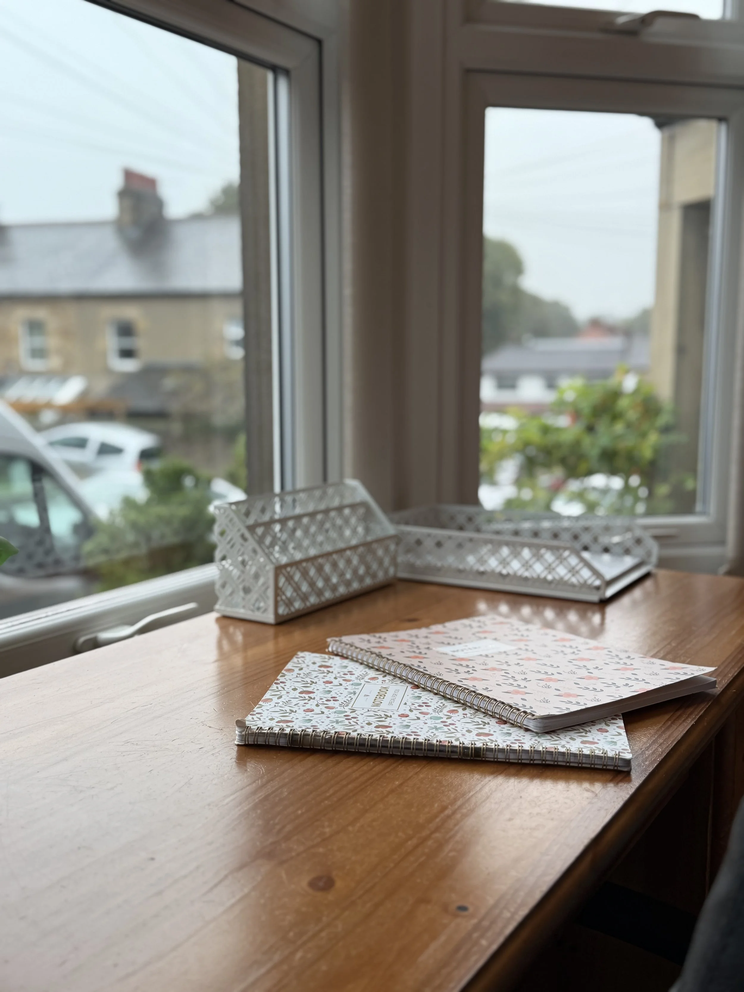 A wooden table near a window with two spiral notebooks with floral covers and two white plastic trays.