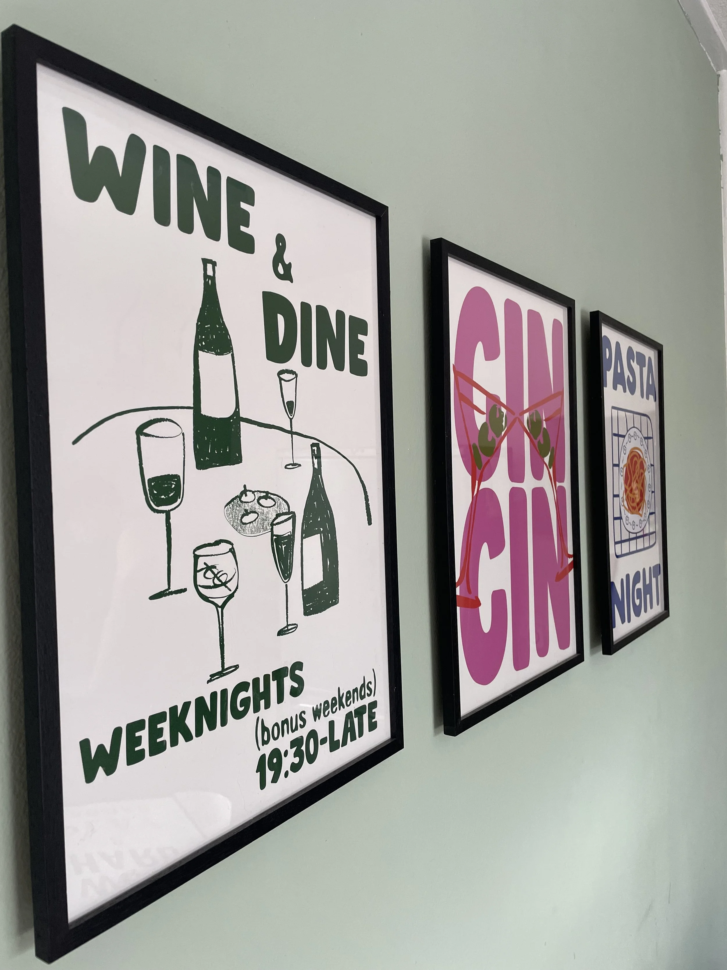 Framed posters on a wall advertising wine, dining, and pasta night with colorful graphics and text.