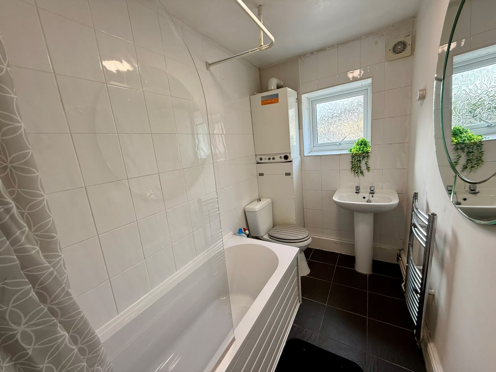A small bathroom with white tiled walls, a bathtub with a patterned shower curtain, a toilet, a small sink with a mirror above it, a heated towel rack, and a small window with frosted glass and two small green plants on the windowsill.