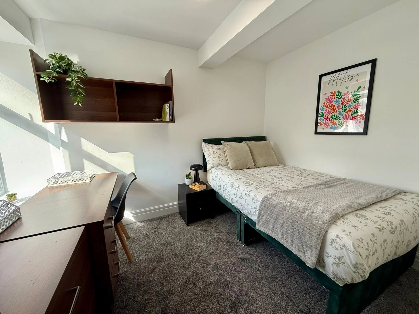A bedroom with a bed, nightstand, desk, and wall shelf, decorated with plants and artwork.