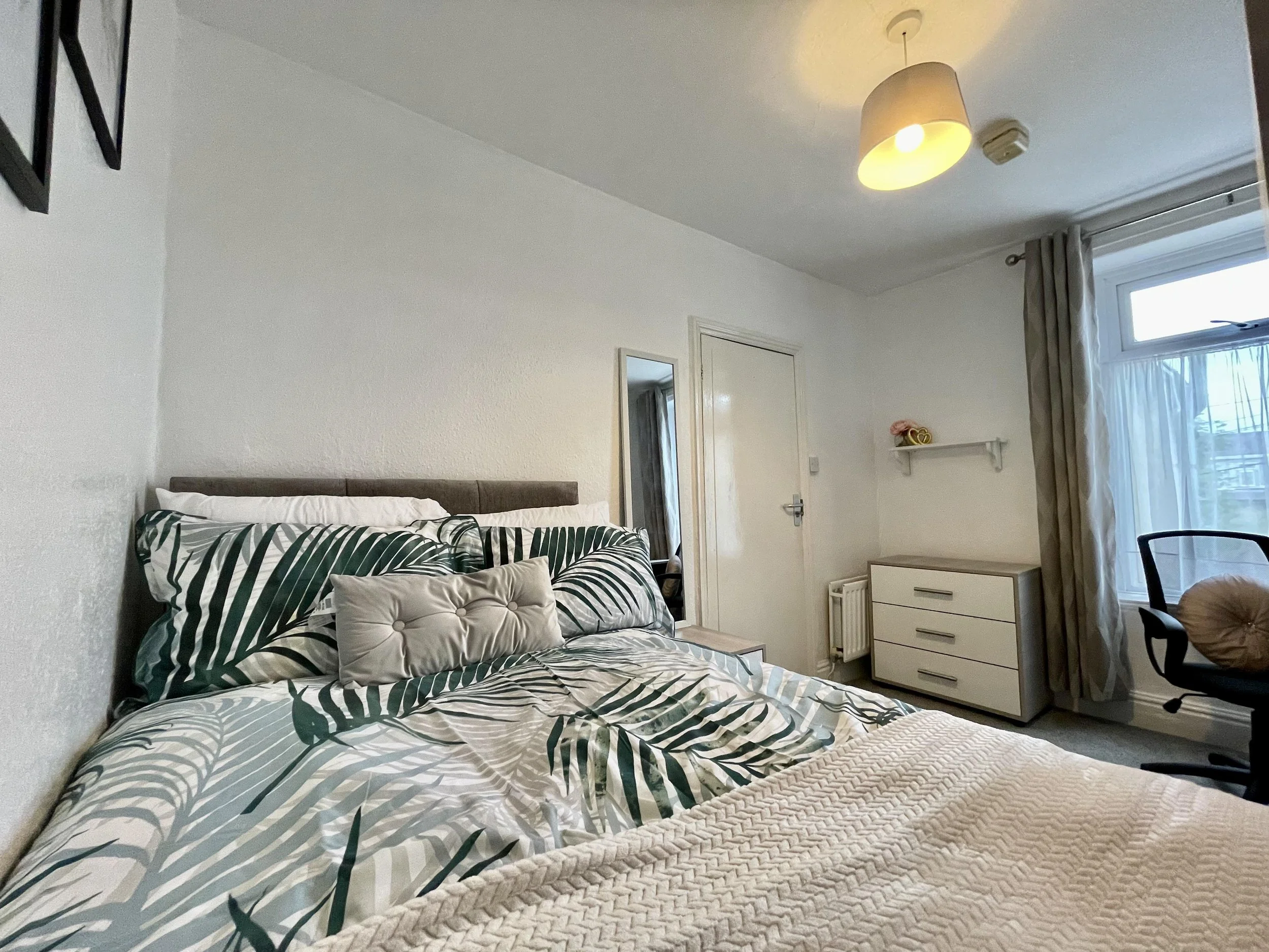 A bedroom with a bed featuring tropical leaf-patterned bedding, a beige tufted pillow, and a window with curtains. There is a chest of drawers, a wall shelf, a chair with a circular cushion, and a ceiling light.