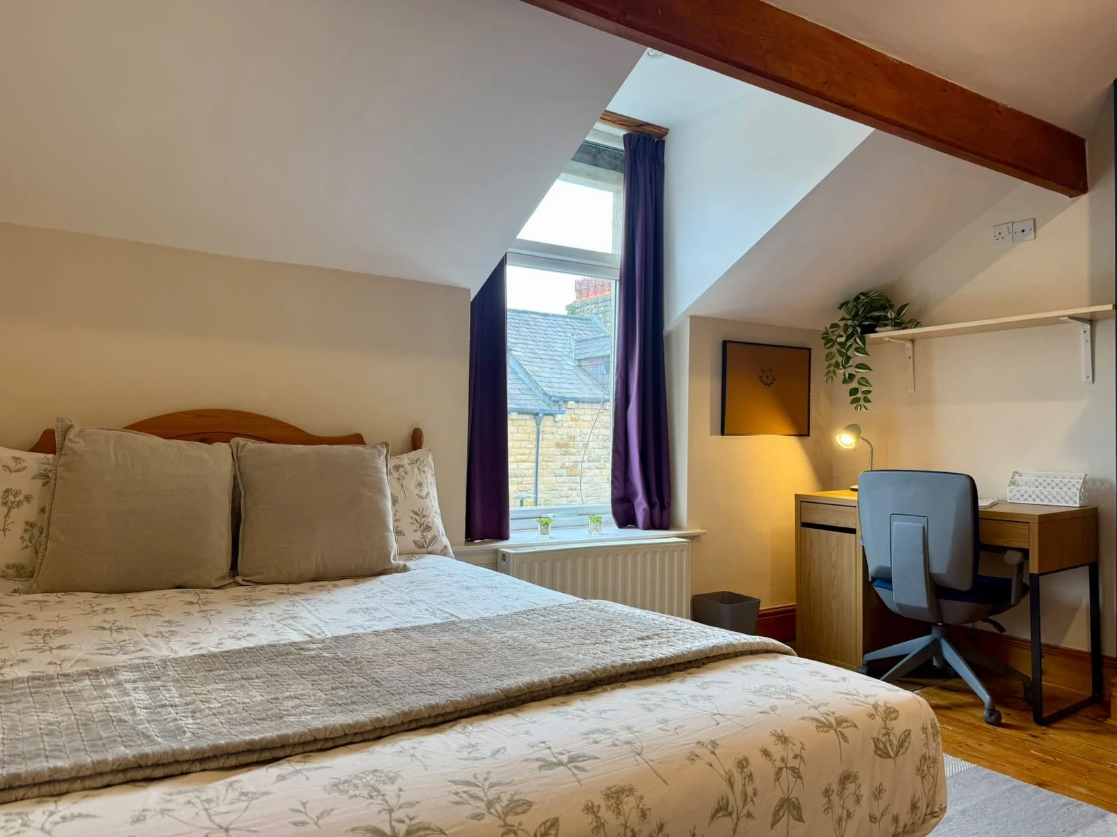 A cozy bedroom with a beige bedspread and pillows, a window with purple curtains, a small desk with a lamp and chair, a plant, and a framed picture on the wall.
