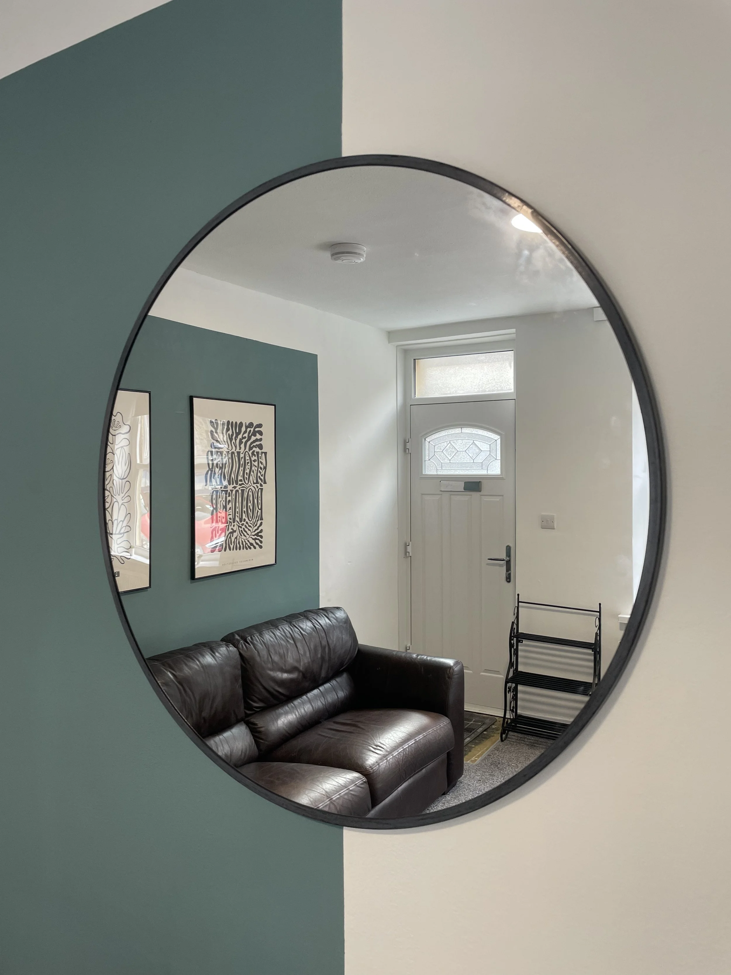 Living room with a black leather sofa, framed art on a teal wall, a white front door, and a black metal shelving unit, seen through a circular mirror on a wall.