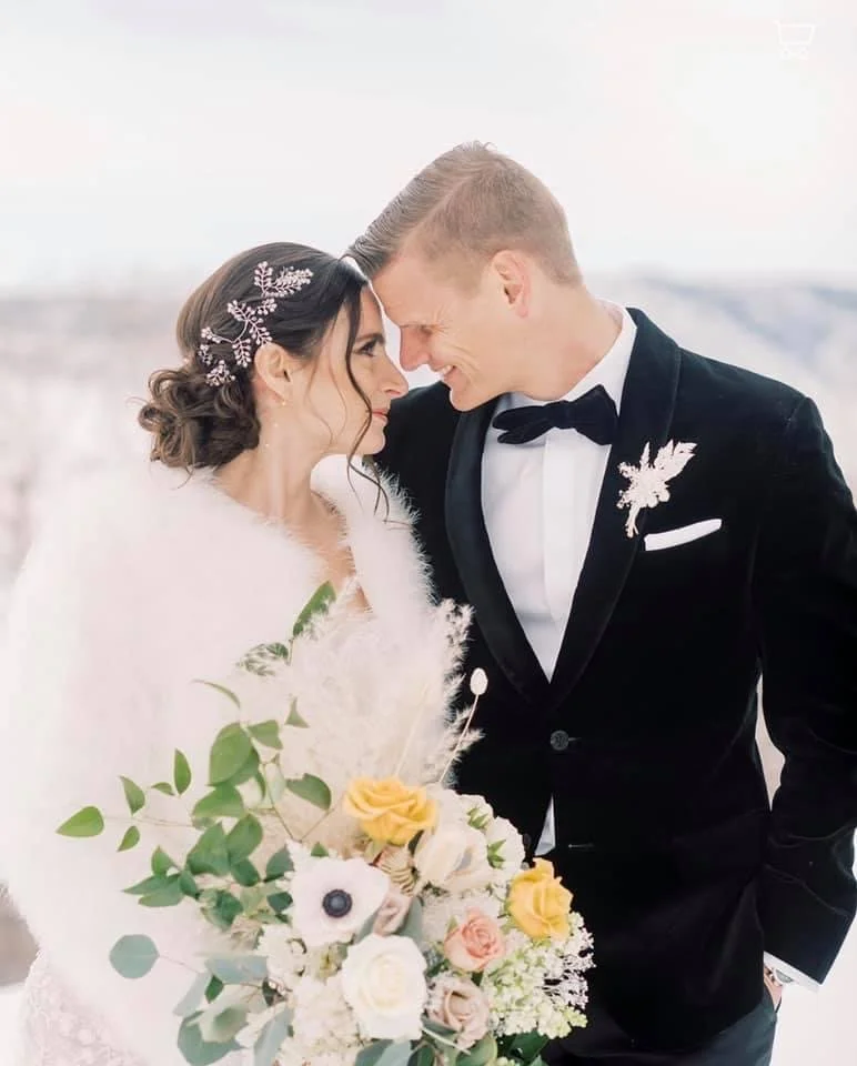 A bride and groom smiling and touching foreheads outdoors during their wedding, with the bride holding a bouquet of flowers.