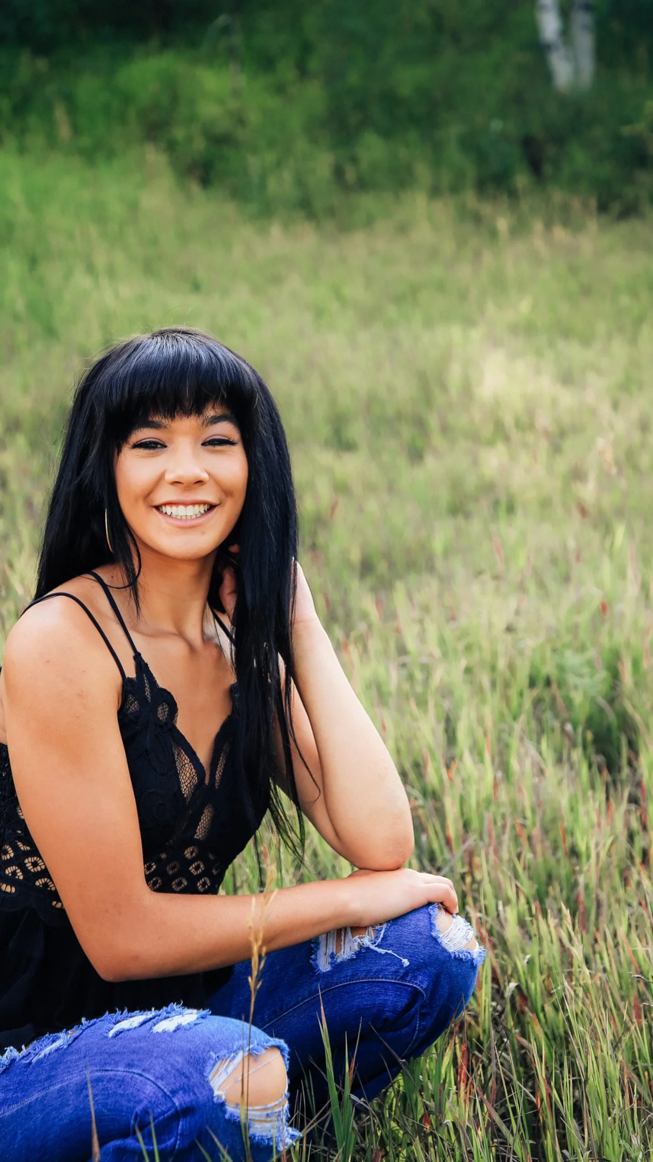 Smiling woman with black hair, wearing a black lace top and ripped blue jeans, sitting in a grassy field.