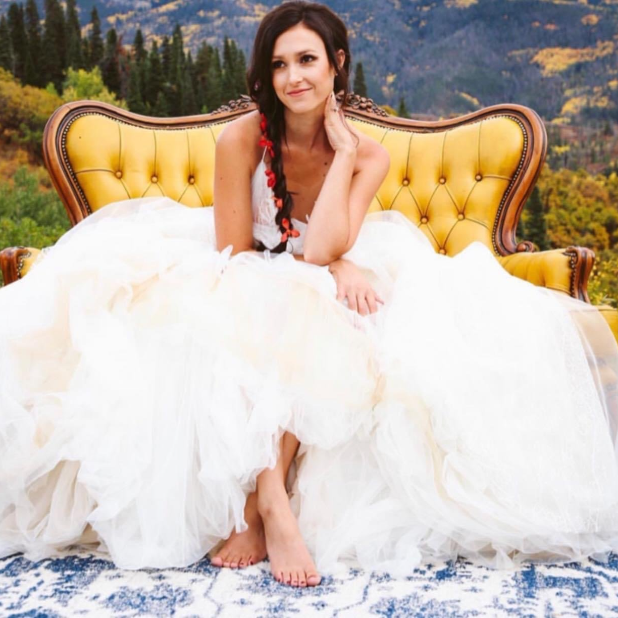 A woman in a white dress sitting on a yellow vintage sofa outdoors with a mountainous forested landscape in the background.