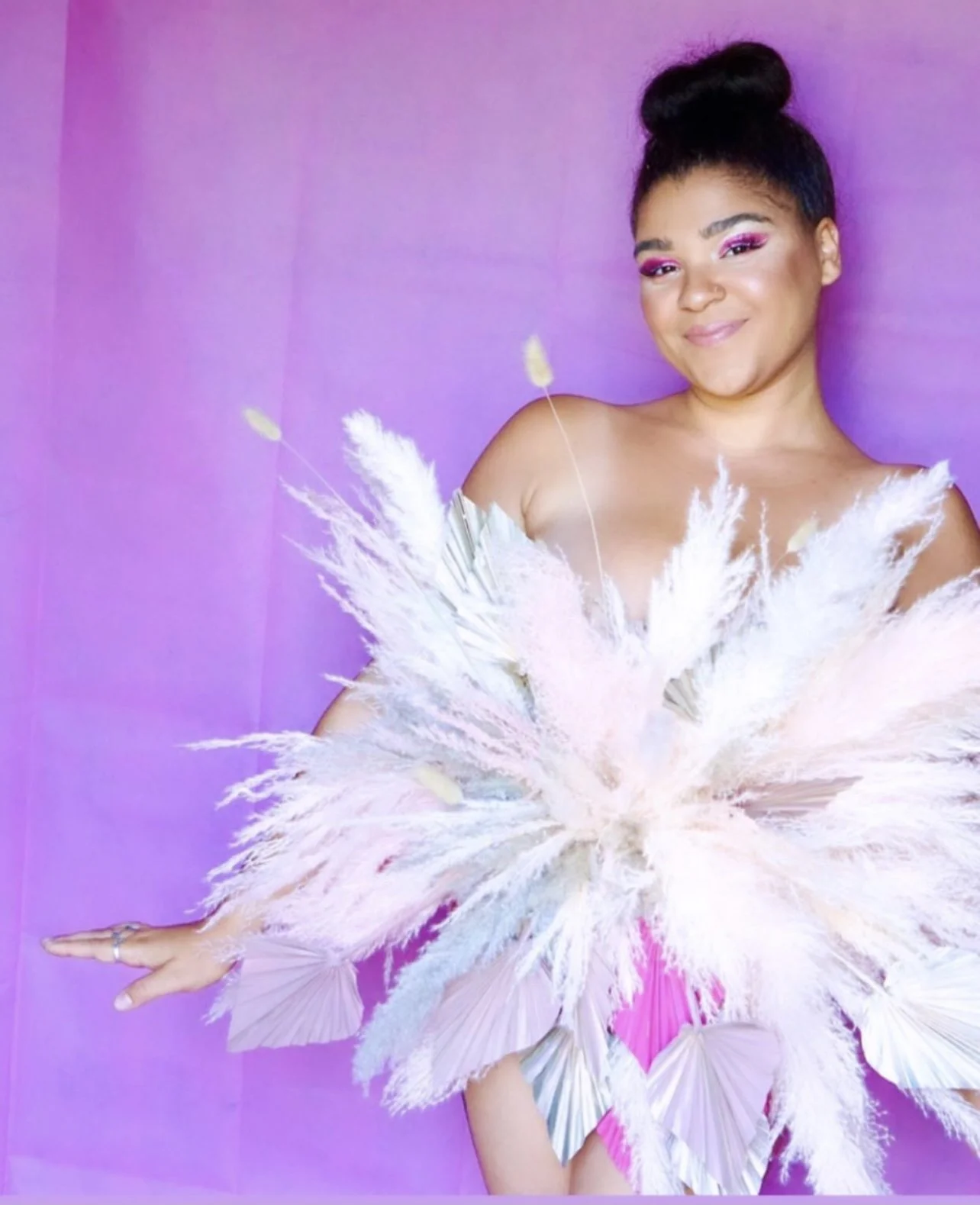 A woman with styled hair and pink makeup, smiling, holds a large bouquet of white and pink feathers and dried flowers, in front of a purple background.