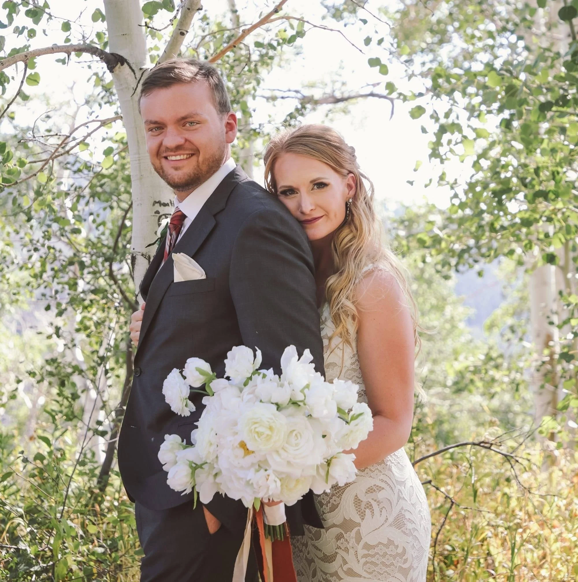 A newlywed couple standing outdoors in a wooded area with trees and greenery, smiling at the camera. The groom is in a black suit with a white shirt, tie, and pocket square, and has his hand in his pocket. The bride is in a lace wedding dress, holding a large white bouquet, with long, wavy blonde hair and earrings.