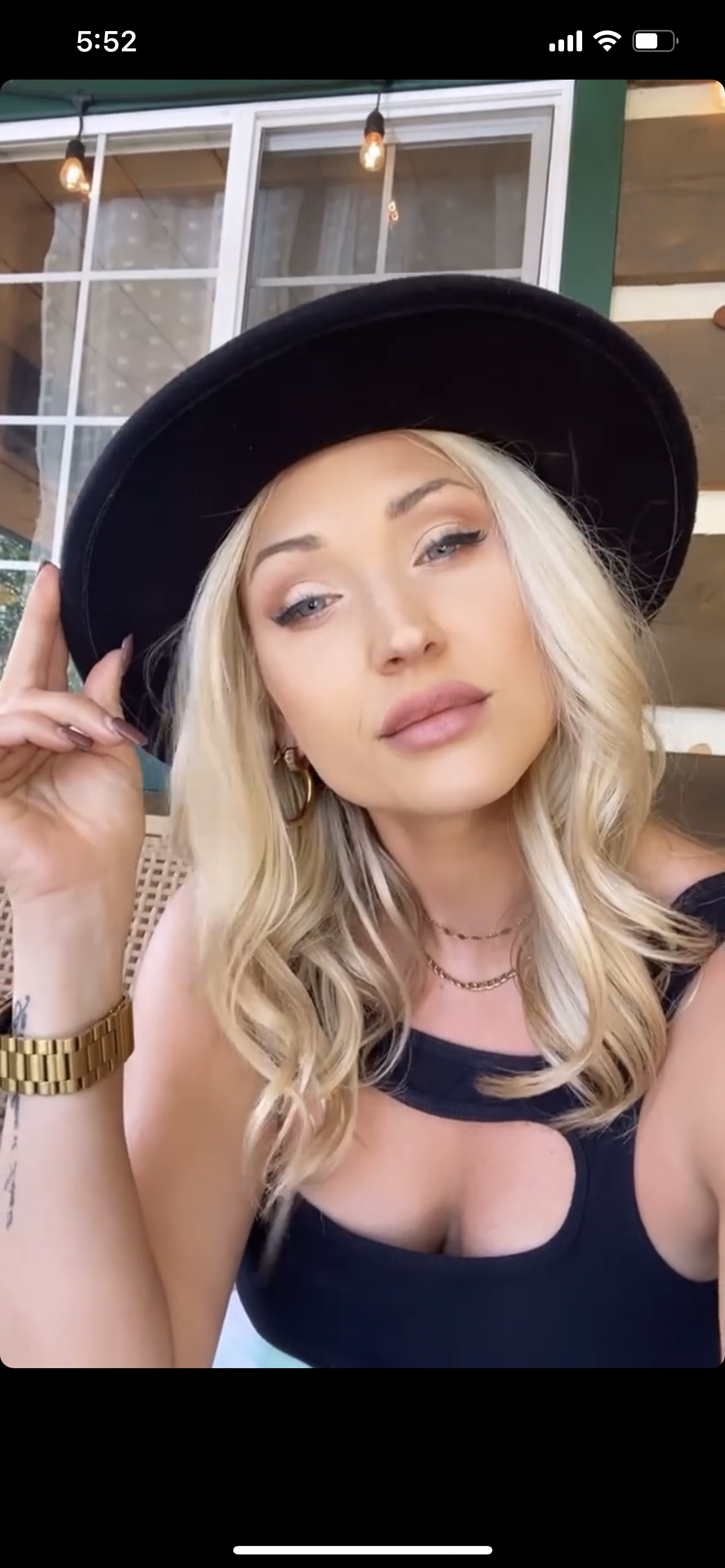 A young woman with blonde wavy hair, wearing makeup, gold jewelry, a black top with a cut-out, a gold watch, and a black wide-brim hat, sitting in an indoor space with large windows and string lights.