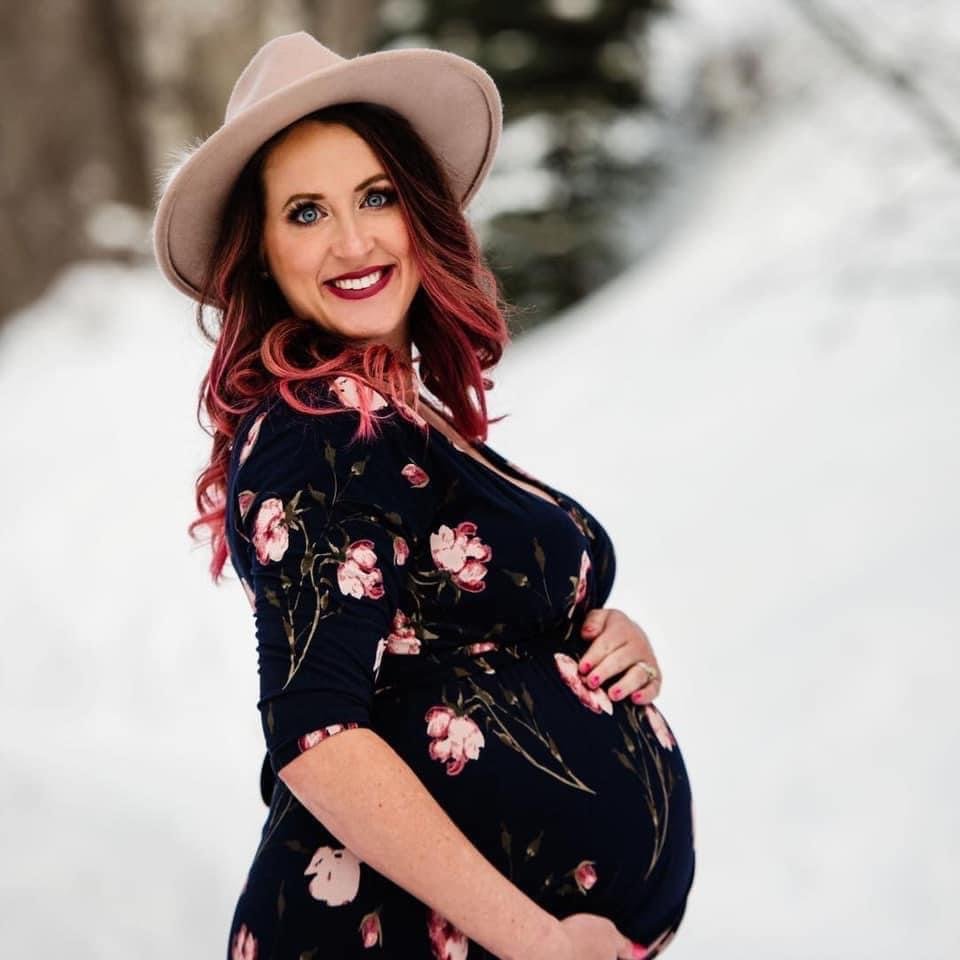 A pregnant woman with red hair, wearing a floral dress and a beige hat, smiling outdoors in a snowy landscape.