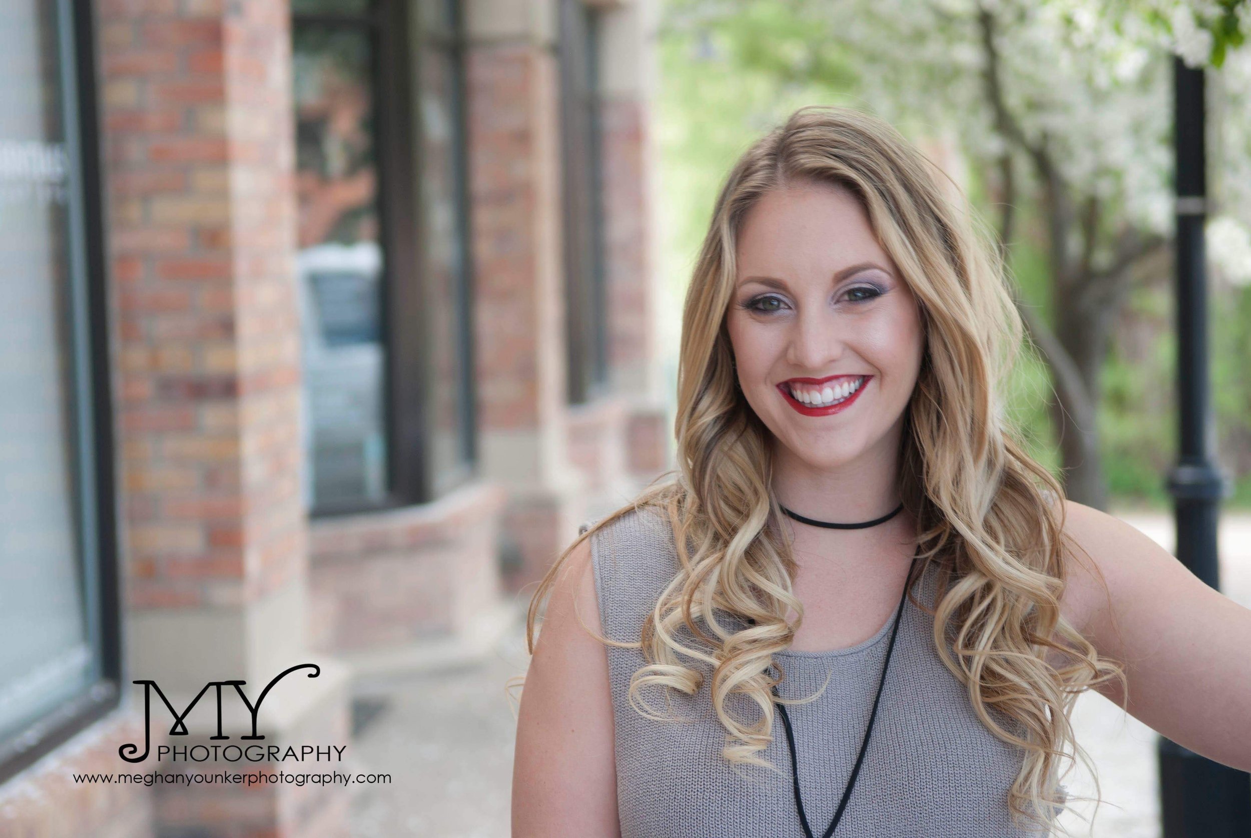 A young woman with long blonde curly hair, bright red lipstick, and makeup, smiling outdoors on a sidewalk with brick buildings and green trees in the background.