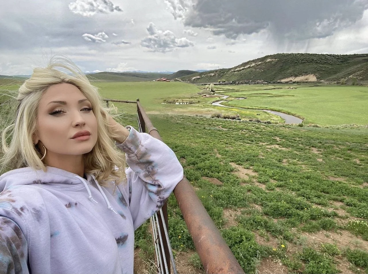 A woman with blonde hair taking a selfie outdoors in a lush green landscape with a winding stream, hills, and cloudy sky in the background.