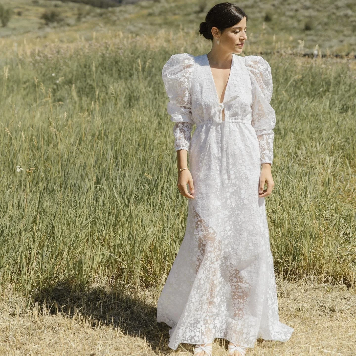 A woman in a white lace dress standing in a grassy field with tall grass and a hill in the background.
