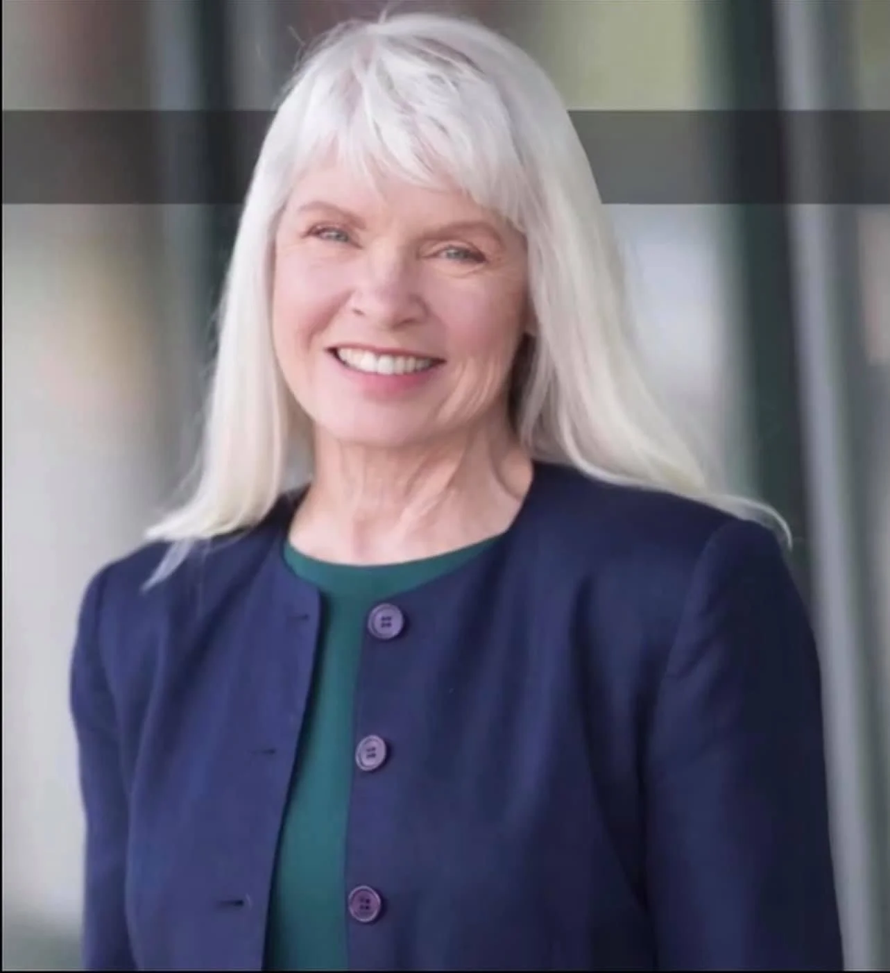 A smiling older woman with long silver hair, wearing a dark blue blazer over a green top, standing outdoors with a blurred background.