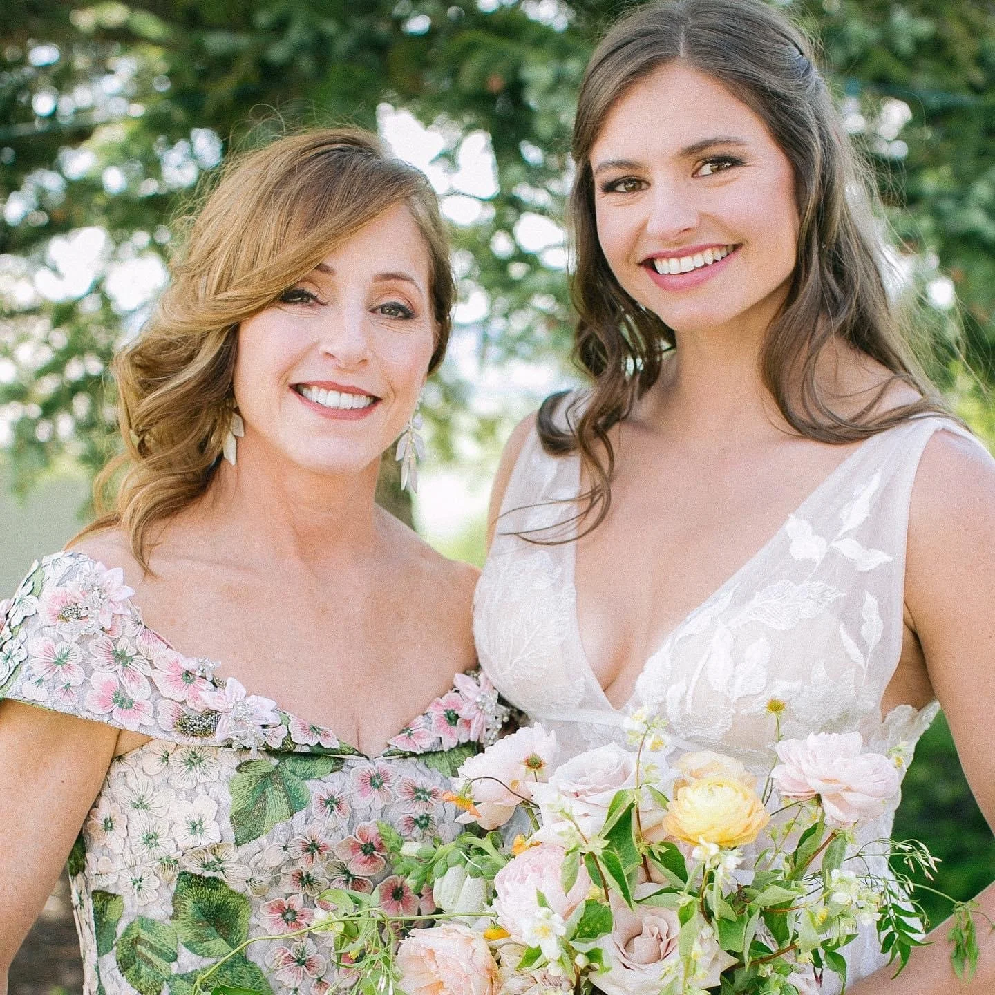 Two women smiling outdoors, one holding a bouquet of flowers, with trees in the background.